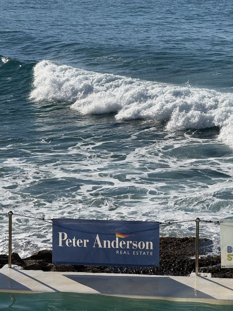 A wave breaks over the Bondi Icebergs pool beside the Peter Anderson Real Estate banner