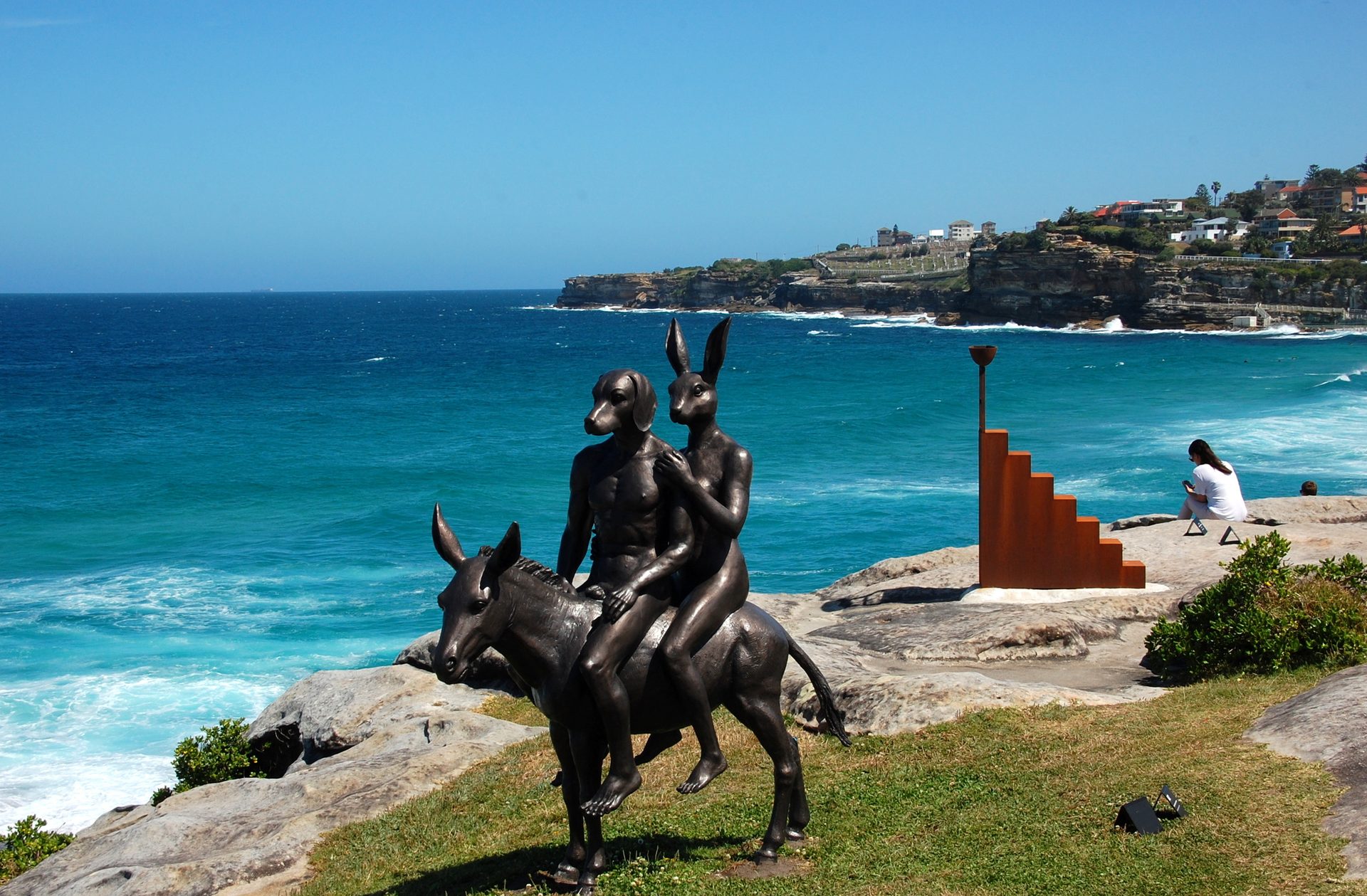 Bronze sculpture of two figures riding a donkey on a grassy headland above the ocean on the Bondi to Tamarama coastal walk